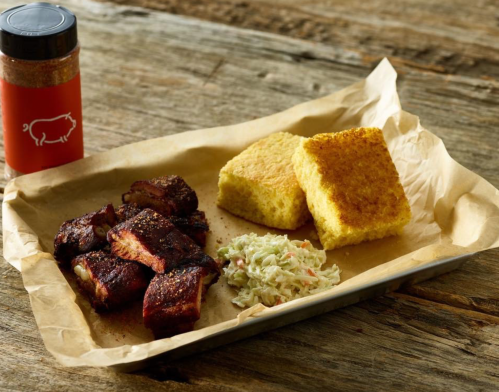 A tray with smoked ribs, coleslaw, and cornbread, alongside a spice jar on a rustic wooden table.