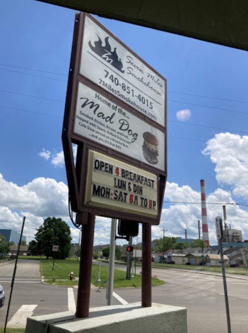 Sign for Seven Miles Smokehouse, advertising breakfast hours and the "Mad Dog" dish, with a blue sky in the background.