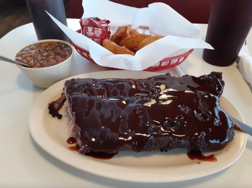 A plate of barbecue ribs covered in sauce, with a side of baked beans and fried items, served with drinks.