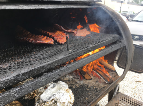 Grill with ribs cooking over an open flame, smoke rising, and foil-wrapped food nearby.