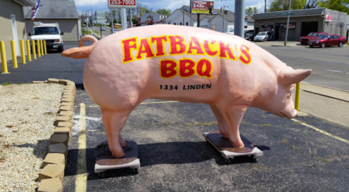 A large pink pig statue with "Fatback's BBQ" written on it, located outside a restaurant on a paved area.