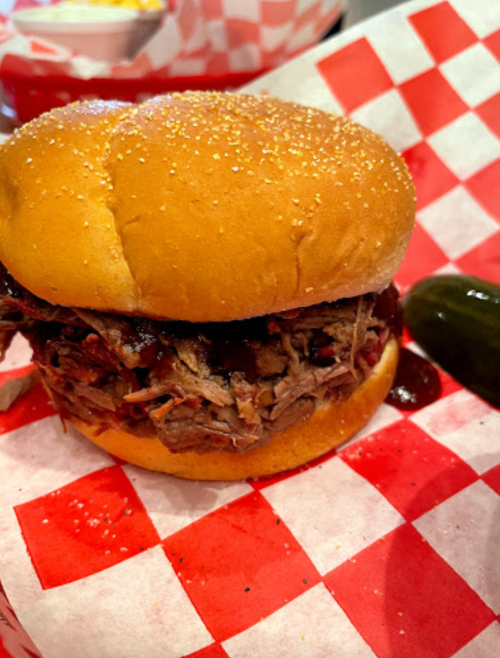 A close-up of a pulled pork sandwich on a sesame seed bun, served with pickles and barbecue sauce on a red-checkered paper.