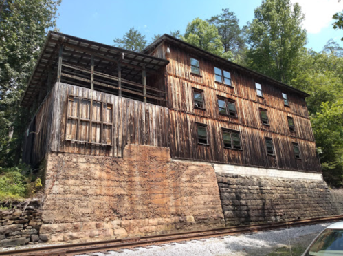 A weathered wooden building with multiple windows, set against a backdrop of trees and a stone foundation.