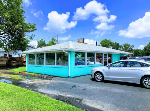 A light blue building with large windows and an "open" sign, surrounded by green grass and parked cars.