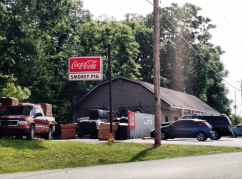 A roadside view of a building with a "Coca-Cola Smokey Pig" sign, surrounded by parked cars and greenery.