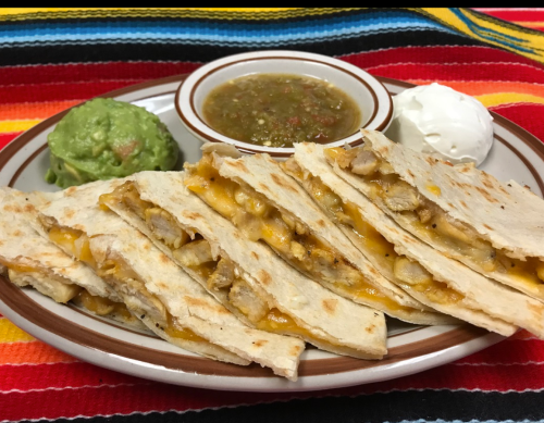 A plate of quesadillas with chicken, cheese, guacamole, salsa, and sour cream, set on a colorful striped background.