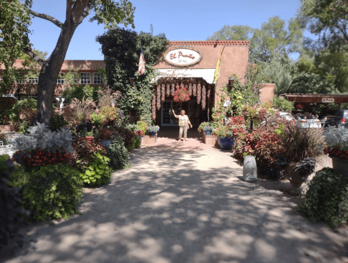 A woman walks towards a restaurant entrance surrounded by vibrant flowers and greenery on a sunny day.