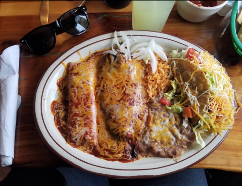 A plate of enchiladas topped with cheese, rice, beans, a taco, and a side salad, with sunglasses nearby.