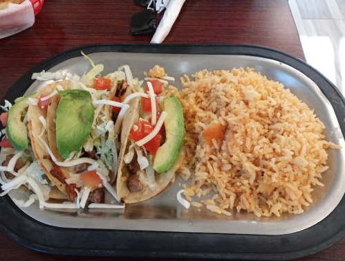 A metal plate with three tacos topped with lettuce, tomatoes, and avocado, alongside a serving of rice.