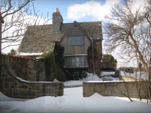 A charming, snow-covered house with a stone wall and trees, set against a cloudy sky.