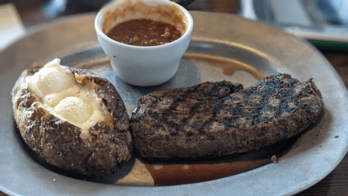 A grilled steak served with a loaded baked potato and a small bowl of chili on a metal plate.