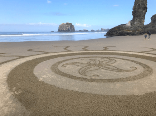 A large sand design resembling a sun on a beach, with rocky formations and ocean waves in the background.
