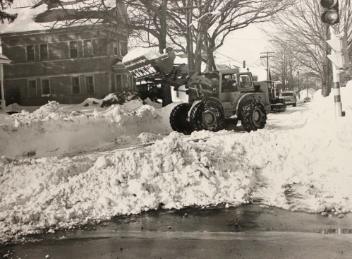 A snowplow clears a snowy street, with large snowbanks and houses in the background on a winter day.
