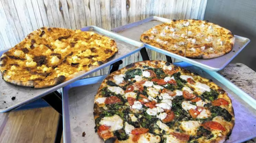 Three pizzas on metal trays: one with cheese, one with spinach and tomatoes, and one with a mix of toppings.