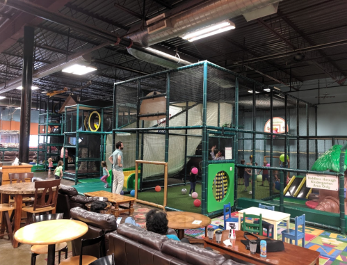 Indoor play area with a large climbing structure, colorful balls, and seating for parents nearby. Kids are playing inside.