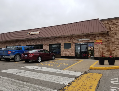 A brick building with a brown roof, featuring a storefront and parked cars in front. Cloudy sky above.