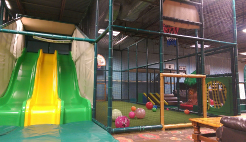 Indoor play area featuring a green slide, basketball hoop, and colorful balls scattered on the turf.