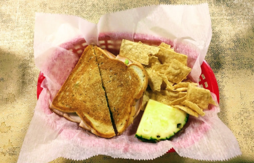 A sandwich with toasted bread, served with tortilla chips and a slice of cucumber in a red basket lined with paper.