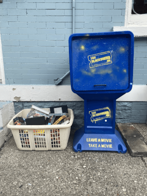 A blue Blockbuster movie rental box next to a basket filled with VHS tapes, labeled "Leave a movie, take a movie."