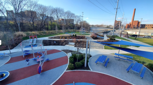 A colorful playground with equipment, picnic tables, and a river view, surrounded by trees and buildings in the background.