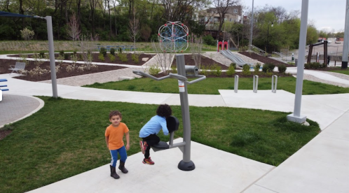 Two children play in a park with exercise equipment and a colorful climbing structure in the background.