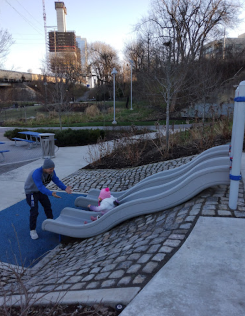 A child slides down a wavy slide while an adult assists them in a park setting with trees and buildings in the background.