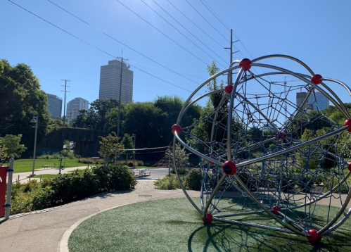 A playground with a large climbing structure, surrounded by greenery and city buildings in the background under a clear blue sky.