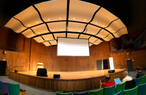A spacious auditorium with a wooden stage, curved ceiling, and empty seats, ready for a presentation.