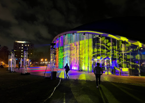 A brightly lit building at night, featuring colorful light projections and people walking outside.