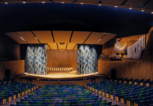 Interior of a concert hall featuring colorful seating, a stage with curtains, and a large pipe organ in the background.