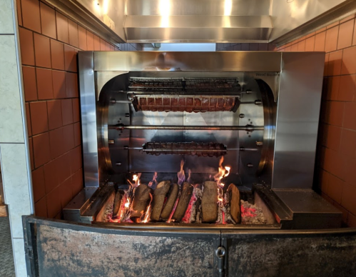A wood-fired grill with meat cooking on a rotating spit over glowing embers.