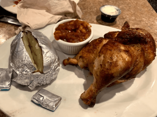 A plate with roasted chicken, a baked potato wrapped in foil, and a small bowl of baked beans.