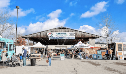 A bustling winter market under a large pavilion, with food trucks, vendor tents, and visitors exploring.