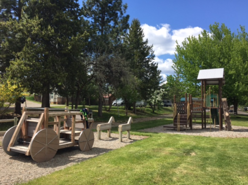 A playground featuring a wooden car structure, benches, and a climbing structure surrounded by trees and blue skies.