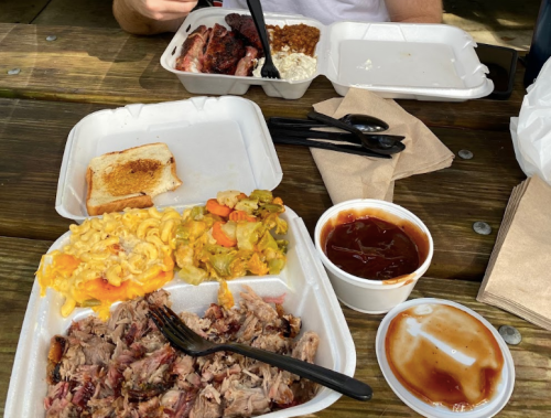 A picnic table with two takeout containers of barbecue, sides, and a slice of toasted bread.