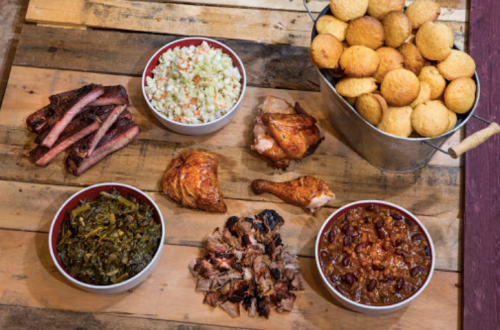 A wooden table displays a variety of barbecue dishes, including ribs, chicken, coleslaw, cornbread, greens, and baked beans.
