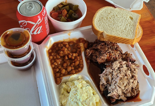 A takeout meal featuring pulled pork, baked beans, potato salad, a side of vegetables, bread, and a can of cola.
