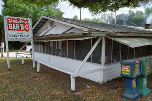 A rustic barbecue restaurant with a sign reading "Peebles Bar-B-Q," featuring outdoor seating and a vintage vending machine.