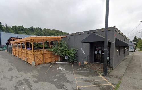 Exterior view of a restaurant with a wooden patio, gray building, and cloudy sky in the background.
