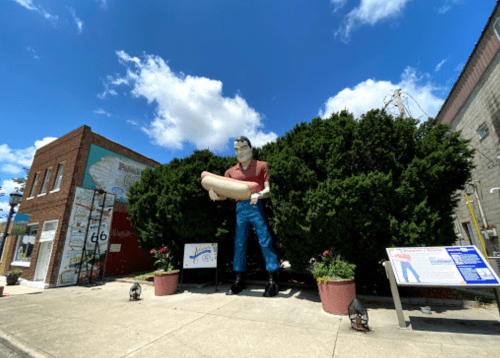 A large statue of a man holding a hot dog stands in front of a green bush and a brick building under a blue sky.