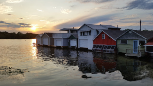 Colorful boathouses on a calm lake at sunset, with reflections in the water and a cloudy sky.