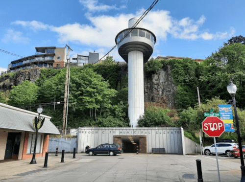 A tall observation tower stands on a rocky hillside, with a stop sign and parked car in the foreground.