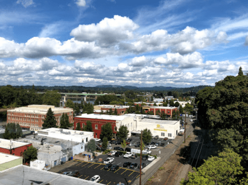 Aerial view of a small town with buildings, parking lots, and mountains in the background under a cloudy sky.