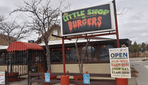 Sign for "Little Shop of Burgers" with a red and black design, featuring outdoor seating and a schedule posted nearby.