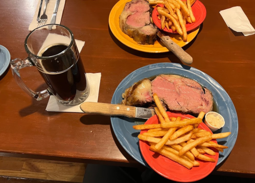 A plate with prime rib, fries, and a side of sauce, alongside a large mug of dark beer on a wooden table.