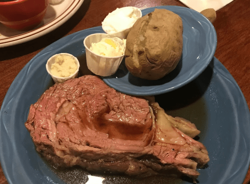 A plate with a juicy prime rib, a baked potato, and two cups of butter and sour cream on a blue dish.