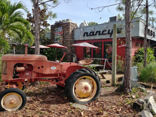 A vintage red tractor sits in front of a colorful restaurant named "Nancy's," surrounded by palm trees and outdoor seating.