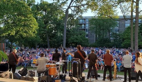 A band performs on stage in front of a large crowd in a park, surrounded by trees and buildings.