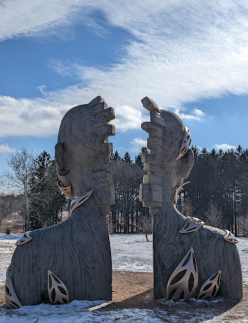 Two large, abstract sculptures of heads with intricate designs, set against a blue sky and snowy landscape.