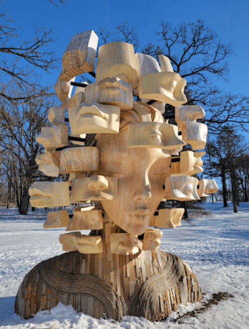 A wooden sculpture of a woman's head with multiple carved faces emerging from her hair, set in a snowy landscape.